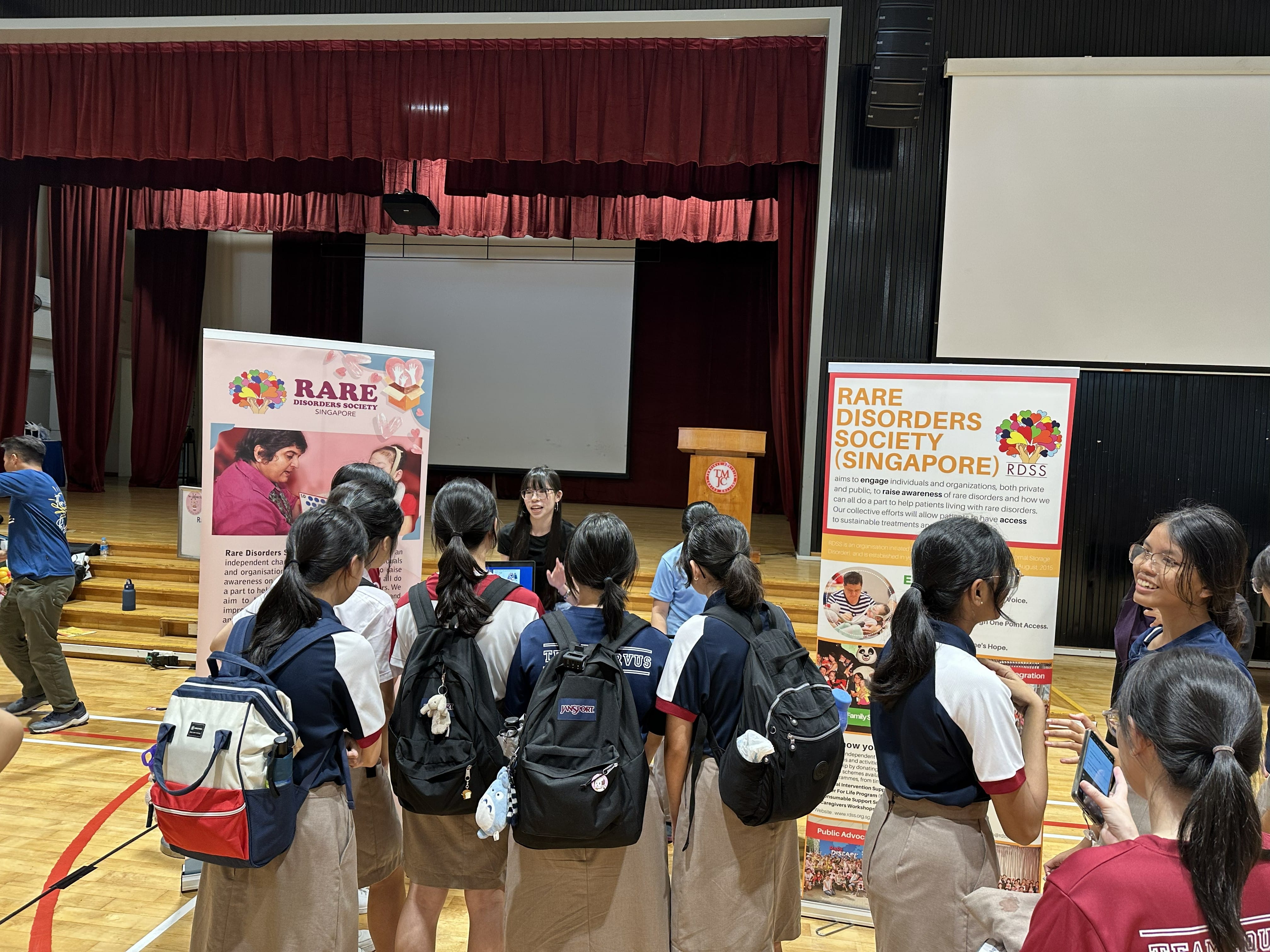 Students in school uniforms gather around exhibition booths for the Rare Disorders Society Singapore (RDSS) inside a school hall. A student presenter engages the crowd in front of two large RDSS banner displays.